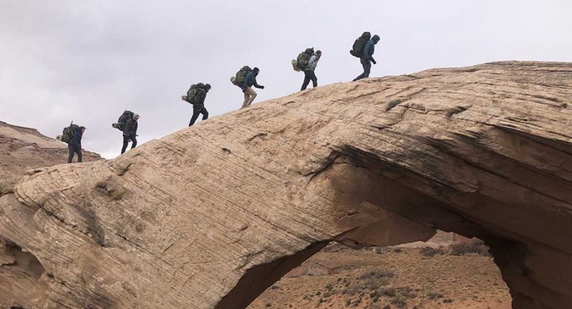 a group of gap year students hike in a line over a rock arch in the american southwest